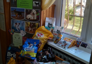 Tablica z obrazkami psów informująca o akcji zbierania karmy. Poniżej kosze z przyniesioną karmą dla psów i kotów w postaci puszek i worków.